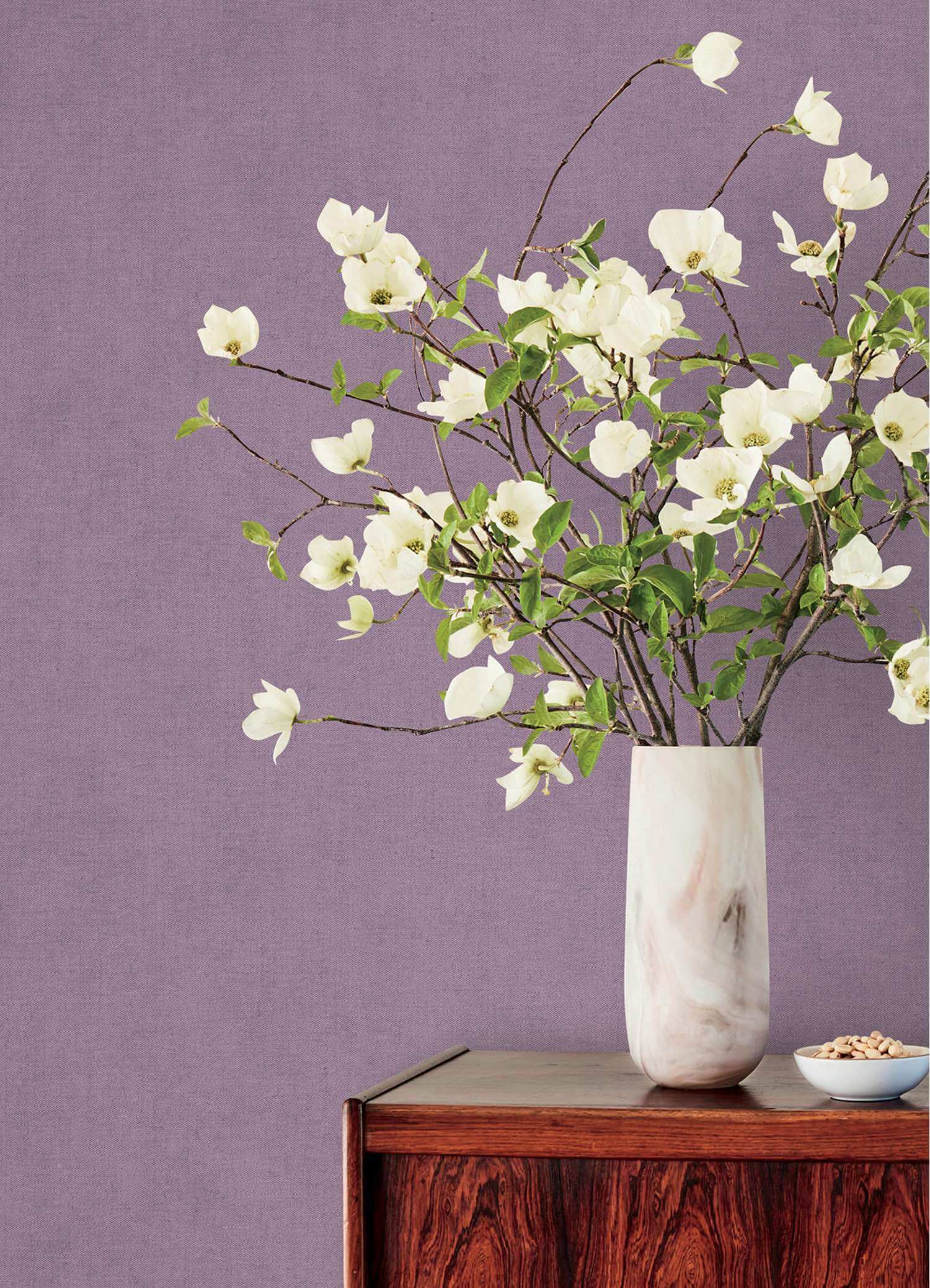 White magnolia flower branches with green leaves arranged in a tall white marble vase on a dark wood sideboard against a textured lavender-purple wall.